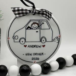 May include: A round white ceramic ornament featuring a black line drawing of a car with a driver waving. The text reads ANDREW, NEW DRIVER, and 2026, accented by small red hearts. A black and white buffalo check ribbon is tied at the top.
