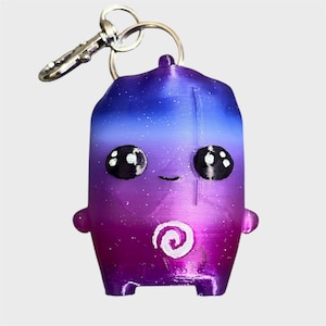 May include: A 3D printed keyring featuring a small, rounded character with a gradient finish transitioning from blue at the top to purple and magenta at the bottom. The figure has large black eyes with white highlights, a small smile, and a white spiral.