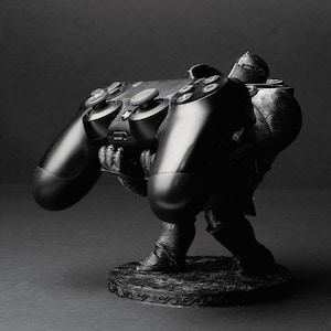 May include: A black and grey 3D printed figurine of a person holding a black PlayStation controller. The figurine is standing on a round base with a decorative design.
