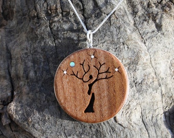 May include: A silver chain necklace with a round wooden pendant. The pendant features a tree design with a person silhouette and small inlaid gemstones. The wood has a warm brown tone, and the necklace is displayed on a textured wooden surface.