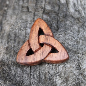 May include: A wooden Celtic knot pendant with a triangular shape. The pendant is made of a light brown wood with visible grain and features a three-part interwoven design. The background is a textured, gray wooden surface.
