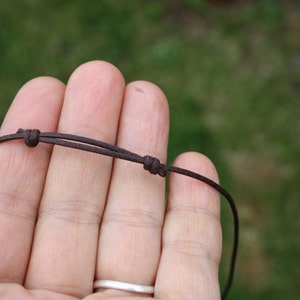 May include: A close-up of a brown cord necklace with a simple knot. The cord is held in a hand, with the background blurred.