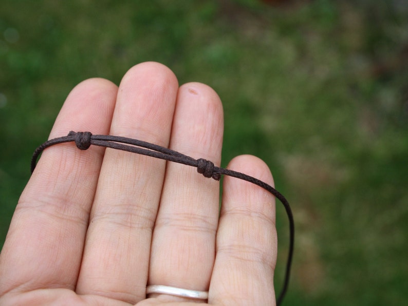 May include: A close-up of a brown cord necklace with a simple knot. The cord is held in a hand, with the background blurred.