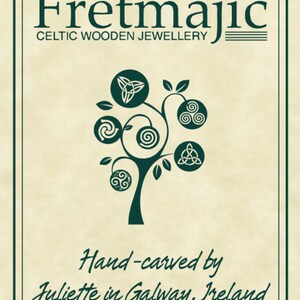 May include: Cream-colored sign with a dark green border featuring the text "Fretmajic Celtic Wooden Jewellery." A stylized tree with Celtic knot designs is in the center. The text "Hand-carved by Juliette in Galway, Ireland" is at the bottom.