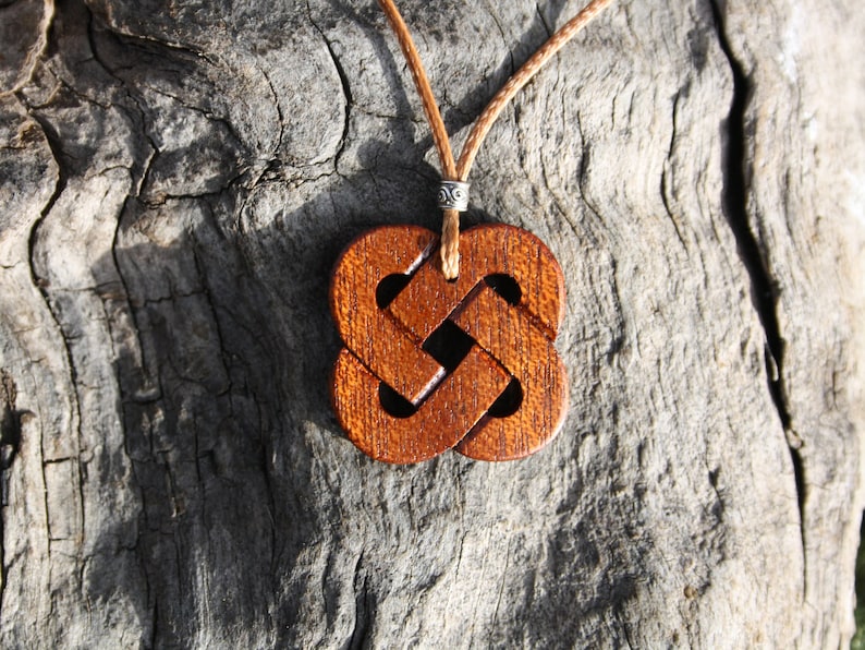 May include: A wooden pendant necklace with a brown Celtic knot design. The pendant is hanging on a brown leather cord.