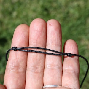 May include: A close-up of a black cord necklace with adjustable knots. The cord is thin and appears to be made of a woven material. The background is blurred, with green grass visible.