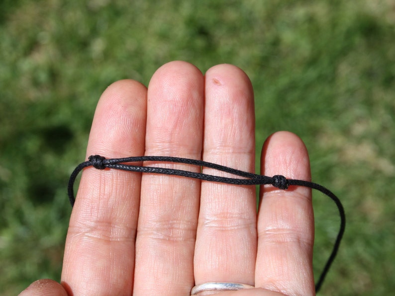 May include: A close-up of a black cord necklace with adjustable knots. The cord is thin and appears to be made of a woven material. The background is blurred, with green grass visible.