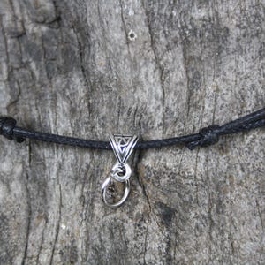 Puede incluir: Un collar de cordón negro con un colgante plateado. El colgante tiene un diseño triangular y un pequeño anillo. El collar está anudado a ambos lados del colgante.