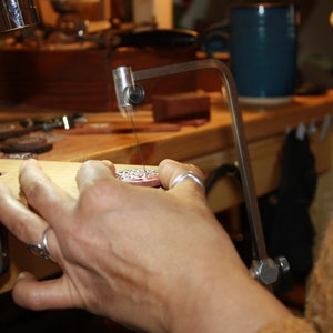 May include: A person's hand holds a silver metal pendant with intricate designs, while using a jeweler's saw to cut a piece of wood.