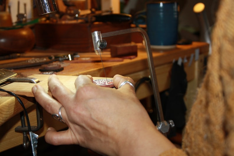 May include: A person's hand holds a silver metal pendant with intricate designs, while using a jeweler's saw to cut a piece of wood.