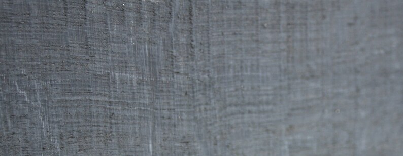 May include: Close-up of a textured, gray surface with visible grain and subtle variations in tone. The surface appears to be made of a rough material, possibly wood or a similar composite, with a slightly weathered appearance.