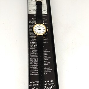Könnte beinhalten: Eine Vintage Buler Royal Armbanduhr mit schwarzem Lederarmband und goldfarbenem Gehäuse. Das Zifferblatt ist weiß mit schwarzen römischen Ziffern und goldenen Zeigern. Die Uhr wird in ihrer originalen schwarzen Box präsentiert, die Text auf Deutsch enthält.