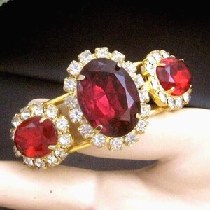 May include: A gold tone bracelet with three large red oval-shaped stones surrounded by clear rhinestones.