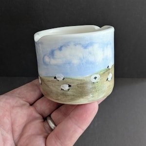 May include: A small ceramic creamer jug featuring a hand-painted landscape scene. The exterior displays a light blue sky with white clouds above a tan grassy field populated by several small white sheep with black faces and legs.