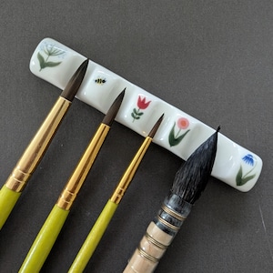 May include: A white ceramic brush rest featuring small painted illustrations of a green plant, a bee, a red tulip, a pink flower, and a blue flower. Four paint brushes with dark bristles and yellow or light wood handles are positioned across the rest.