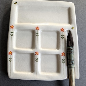 May include: A white ceramic watercolour palette with five square mixing wells and one large rectangular well. The palette features hand-painted orange flowers with green stems and small yellow and black bees. A wooden paintbrush rests on the side.