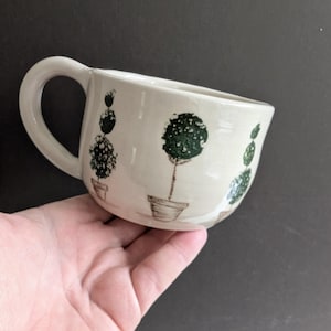 May include: Cream ceramic mug with a rounded shape and a large handle. The mug is decorated with hand-painted green topiary designs in brown pots. The mug is held in a hand against a dark background.
