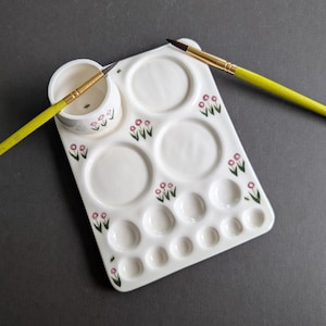 May include: A white ceramic artist's palette with a built-in paint cup and multiple wells for mixing colours. The palette is decorated with pink floral designs and small bees. Two paintbrushes with yellow handles and gold ferrules are placed on the palette.