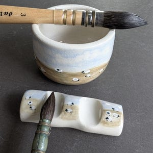 May include: A ceramic water bowl and a matching brush rest, both featuring a hand-painted landscape with small white sheep on a tan field under a light blue sky. Two dark-bristled calligraphy brushes with wooden handles are positioned on the items.