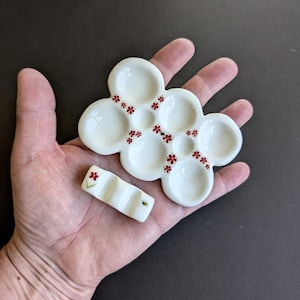 May include: A white ceramic paint palette with multiple wells and a matching brush rest. Both pieces are decorated with red flower accents. A small bee is painted on the brush rest. The palette is held in a hand.