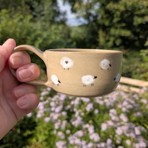 May include: A beige ceramic mug with a smooth finish, featuring a repeating pattern of small, hand-painted white sheep with black details. The mug has a single curved handle on the side and a wide, cylindrical body with a flat base.