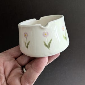 May include: A small, off-white ceramic ashtray featuring a hand-painted pattern of delicate pink flowers with yellow centres and green stems. The bowl has a smooth, glossy finish and includes two distinct notches along the top rim for resting items.