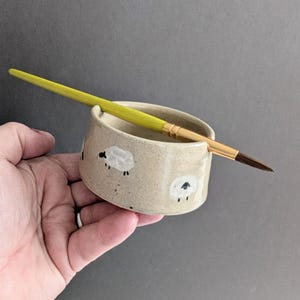 May include: A beige ceramic brush holder with a sheep design. The holder has a rounded shape and is speckled with black dots. A yellow and gold paintbrush rests in the holder. The holder is held in a hand.