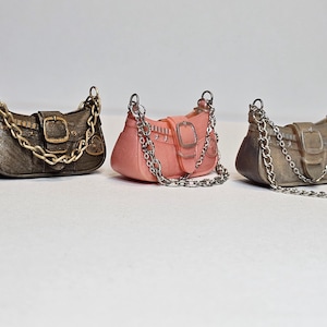Könnte beinhalten: Drei Miniaturhandtaschen in Metallic-Bronze, Rosa und Grau. Jede Tasche hat einen Kettenriemen und einen Schnallenverschluss. Die Taschen werden auf einer weißen Oberfläche präsentiert.