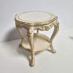 Könnte beinhalten: Kleiner, verzierter, weißer Beistelltisch mit goldenen Akzenten. Der Tisch hat eine runde Oberseite und ein unteres Regal, beide mit strukturierter Oberfläche. Die geschwungenen Beine und dekorativen Elemente verleihen ihm einen Vintage-Look.
