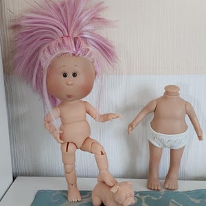 Könnte beinhalten: Eine Puppe mit rosa Perücke und beweglichen Gliedmaßen steht mit einem Fuß auf einer kleinen rosa Mopsfigur. Ein weiterer Puppenkörper mit weißer Unterwäsche befindet sich in der Nähe. Die Puppen stehen auf einer blau gemusterten Oberfläche.