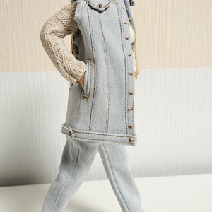 Könnte beinhalten: Puppe mit hellblauer Jeansweste und hellbeigem Strickpullover. Die Puppe trägt außerdem eine passende hellblaue Hose und graue Turnschuhe. Die Puppe hat kurze schwarze Haare und goldene Ohrringe.