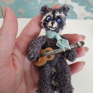 Könnte beinhalten: Eine handgefertigte Miniatur-Waschbärpuppe mit grau-weißem Fellmuster, blauen Augen und einer hellblauen Krawatte. Die Puppe hält eine kleine, braune Gitarre. Die Puppe besteht aus einem flauschigen Material.