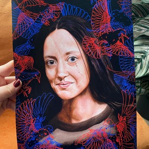 May include: Art print of a woman with long dark hair and a brown top, framed by red and blue outlined birds on a black backdrop. The woman has a scar on her face and is smiling. The print is held by a person.