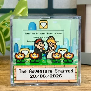 May include: A clear acrylic block with a pixel art wedding scene. The text reads "The Adventure Started 20/06/2026." The image shows a pixelated couple, flowers, and a green landscape, all in a retro video game style.