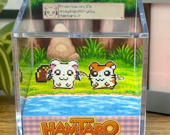 Puede incluir: Una caja de exhibición de acrílico transparente con personajes Hamtaro pixelados en una escena de bosque. La caja incluye el texto "From now on, I'm staying with you, Hamtaro." y el logo de Hamtaro con "Ham-Ham Heartbreak".