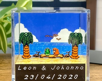 Puede incluir: Bloque acrílico transparente con una escena de playa pixelada. Muestra palmeras, mar azul y dos figuras en un tronco. El bloque tiene los nombres "Leon & Johanna" y la fecha "23/04/2020" en la parte inferior.