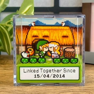 Può includere: Un blocco di acrilico trasparente con una scena pixelata di due personaggi che si baciano, con uno sfondo di una casa e un cielo blu. Il testo "Linked Together Since 15/04/2014" è visualizzato su una base verde. Oggetto decorativo.