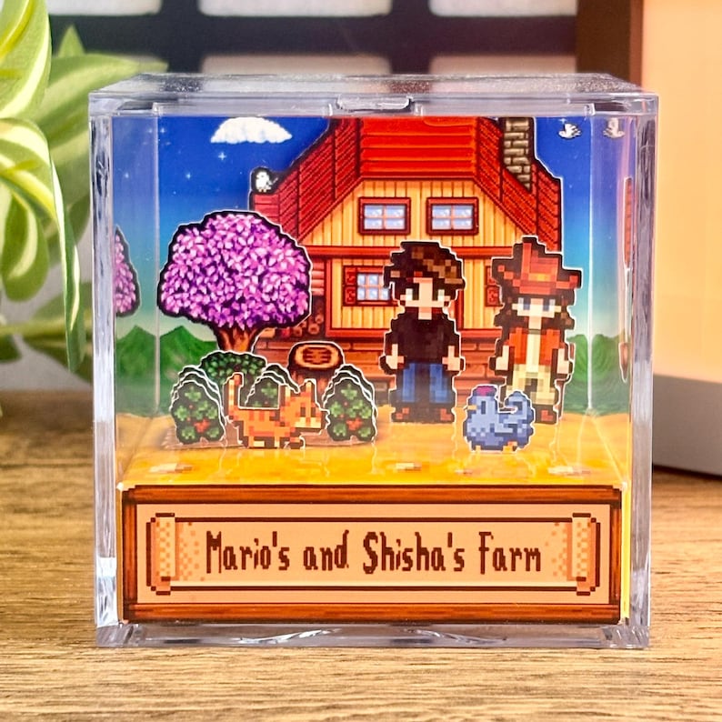 May include: A clear acrylic block featuring a pixelated farm scene. The scene includes a red-roofed house, two figures, a cat, and a blue creature. A purple tree and green bushes are also present. The block has the text "Mario's and Shisha's Farm".