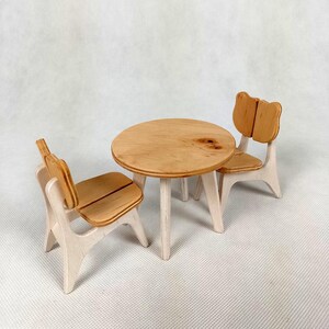Może przedstawiać: Miniaturowy zestaw drewnianego stołu i dwóch krzeseł. Stół jest okrągły z trzema nogami, a krzesła mają oparcie w kształcie misia. Zestaw wykonany jest z jasnego drewna z naturalnym wykończeniem, idealny do domku dla lalek lub zabaw dzieci.