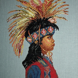 May include: A cross-stitch portrait of a young person wearing a colorful headdress with feathers. The person is looking to the right and has dark hair and brown skin.