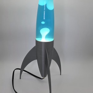 May include: A retro-style lava lamp shaped like a rocket. The lamp has a gray base and fins, a clear glass cylinder filled with blue liquid and white blobs. A black power cord with an on/off switch extends from the base.