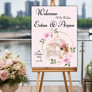 May include: A pink wedding welcome sign with a gold frame and floral accents. The sign features a couple in traditional Indian attire standing in front of a white gazebo. The text on the sign reads "Welcome to Our Wedding Estaa & Aryan 25 October 2024 6:00 PM".