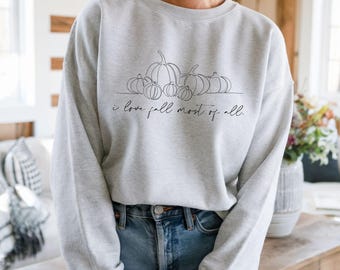 Puede incluir: Una sudadera gris claro con un dibujo lineal de calabazas y el texto "I love fall most of all." La camiseta de manga larga y cuello redondo se combina con unos vaqueros azules. El fondo incluye una mesa de madera y un jarrón con flores.