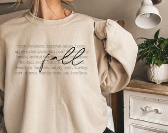 Puede incluir: Una sudadera beige con la palabra "fall" en escritura cursiva. El texto enumera elementos temáticos de otoño como suéteres acogedores, sidra de manzana, campos de calabazas y laberintos de maíz. La sudadera es de color neutro.