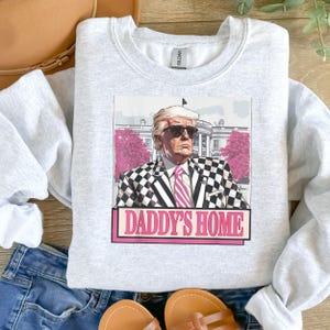 Może przedstawiać: Szara bluza z nadrukiem graficznym przedstawiającym mężczyznę w kratę i okularach przeciwsłonecznych. Tekst "Daddy's Home" jest nadrukowany na białym tle kolorem różowym.