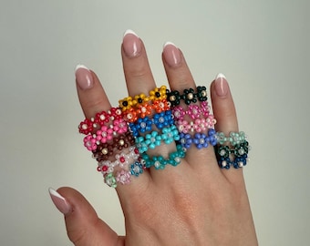 May include: A hand displays a collection of beaded flower rings in various colors. The rings feature small, colorful beads arranged in a daisy-like pattern. Colors include pink, blue, green, yellow, and white. The rings are worn on multiple fingers.