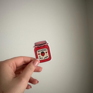 May include: A sticker in the shape of a jam jar with the text "Reading is my jam" and a strawberry illustration. The jar is red with a pink lid and white text. The sticker is held up against a white background.