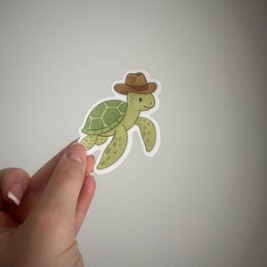 May include: A sticker featuring a cartoon turtle wearing a brown cowboy hat. The turtle has a green shell with a patterned design and is smiling. The sticker is held against a plain white background.