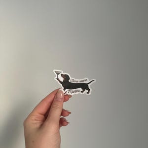 Puede incluir: Una pegatina con un perro salchicha negro sosteniendo una copa de martini. El texto "Teenie weenie Martini" está escrito en cursiva. La pegatina está sobre un fondo gris.