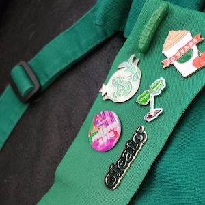May include: A green fabric apron with various pins attached. The pins include a green pickle, a green sprout, a green and white pomegranate, a pink and white circle with the text "Make Some Noise", a gold pin with the text "Oleato", and a white coffee cup with the text "20 Years" and a red cherry.